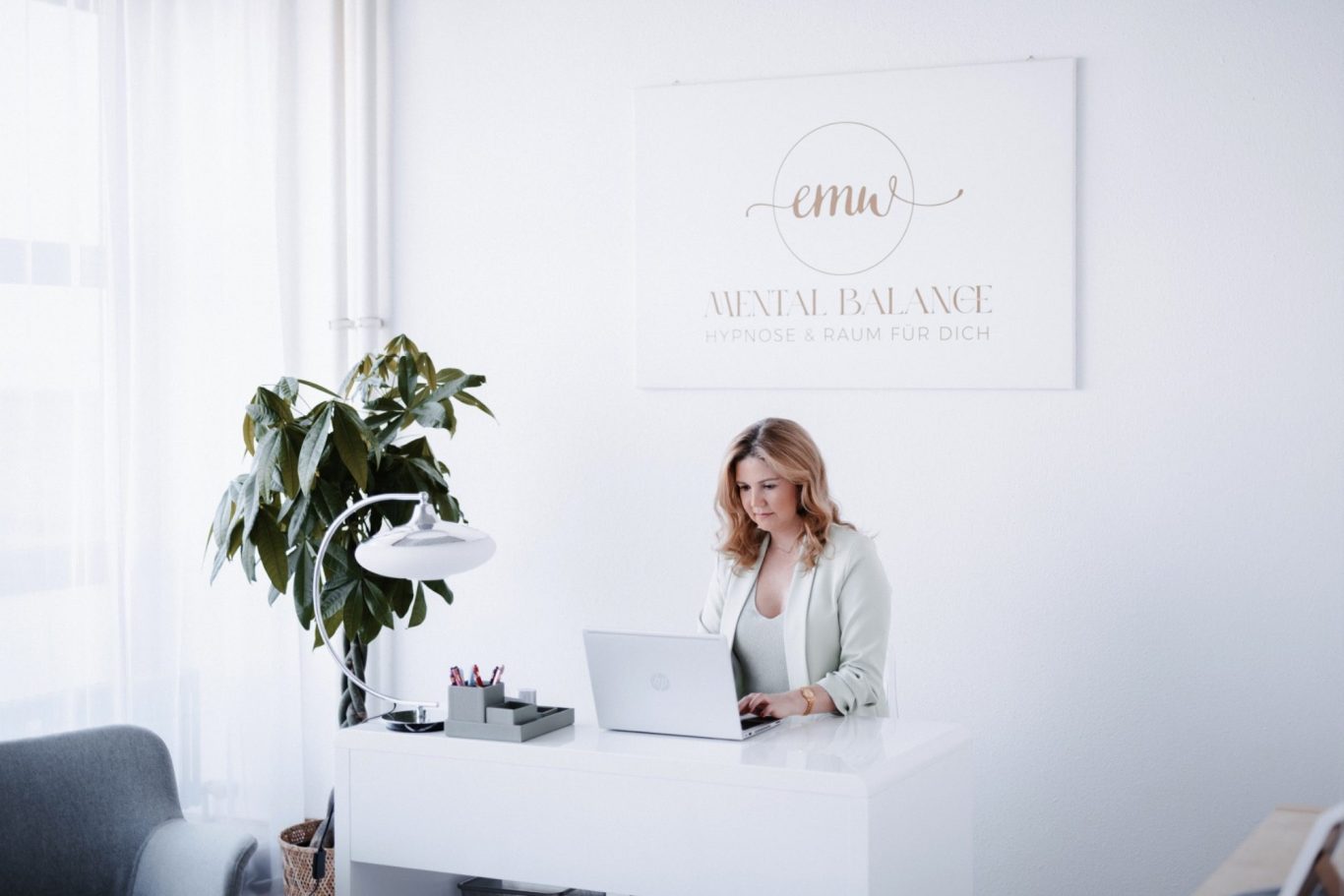 Frau arbeitet am Laptop sitzend am Schreibtisch. Hinter ihr ein Wandbild mit dem Schriftzug „emw - Mental Balance, Hypnose & Raum für Dich“